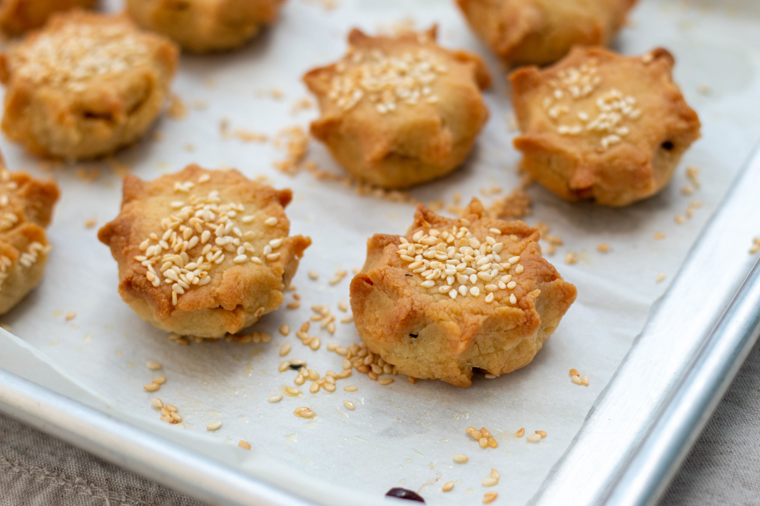 Small savory pastries called pastelicos topped with sesame seeds on a piece of parchment paper
