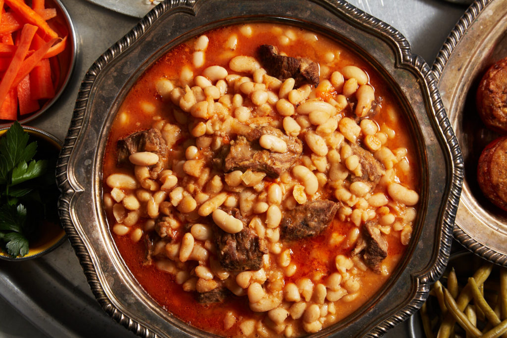 A soup of white beans and meat in a tomato broth in a metal serving bowl