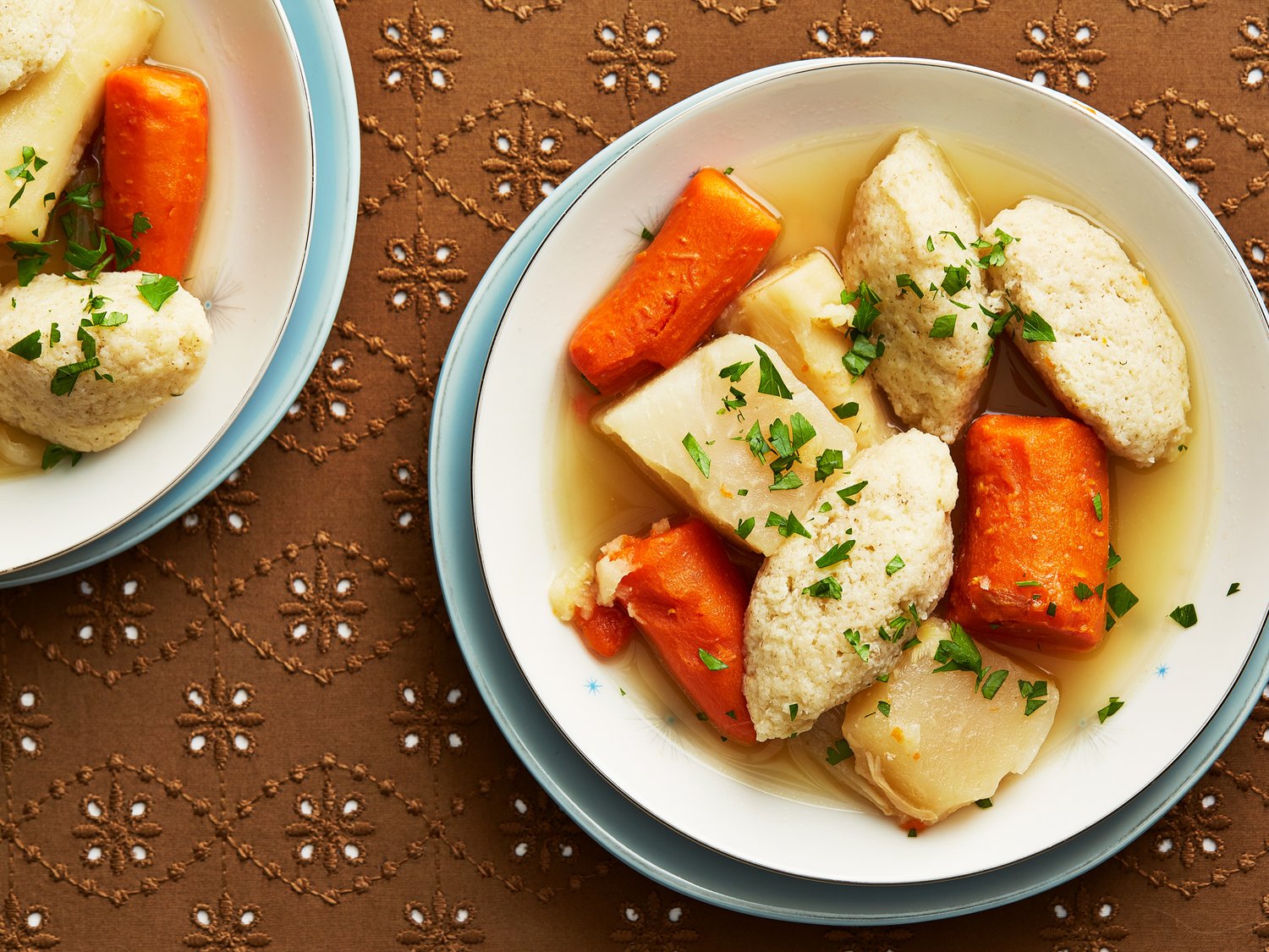 White bowl of chicken soup with dumplings and chunks of root vegetables sits atop brown tablelcoth