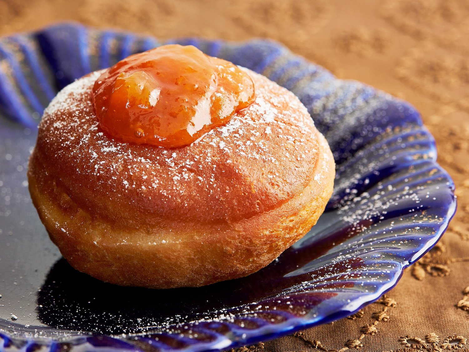 Dome-shaped doughnut topped with apricot jam sits atop blue plate on brown tablecloth