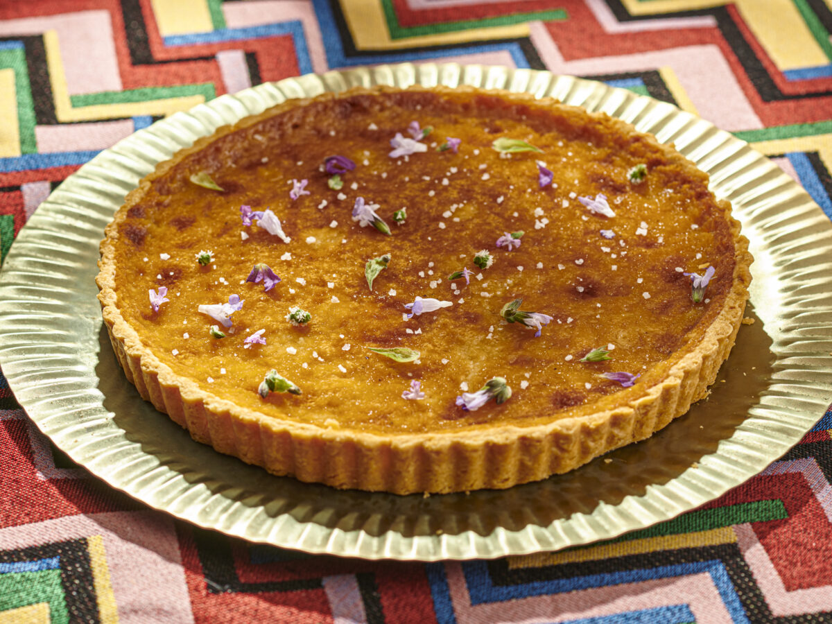 Round and golden honey tart dotted with edible flowers sits atop a gold tray and a colorful tablecloth