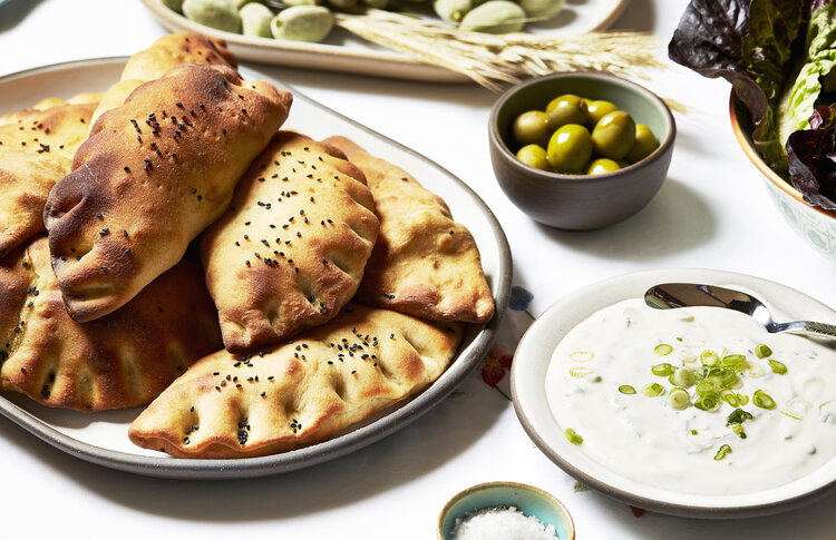 Kurdish stuffed hand pies are served on a white ceramic platter next to bowls of olives and yogurt sauce
