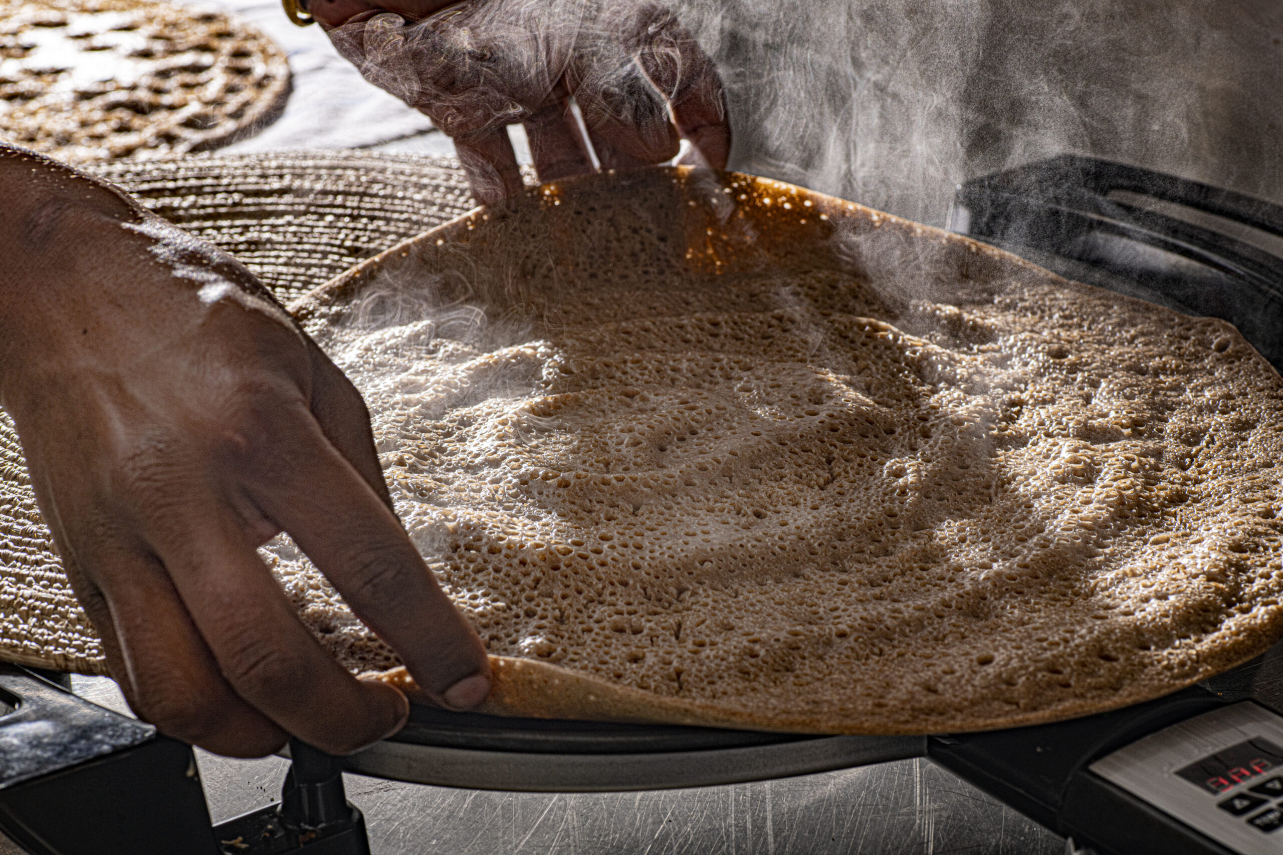 Two brown hands lift a steaming Ethiopian flatbread called injera