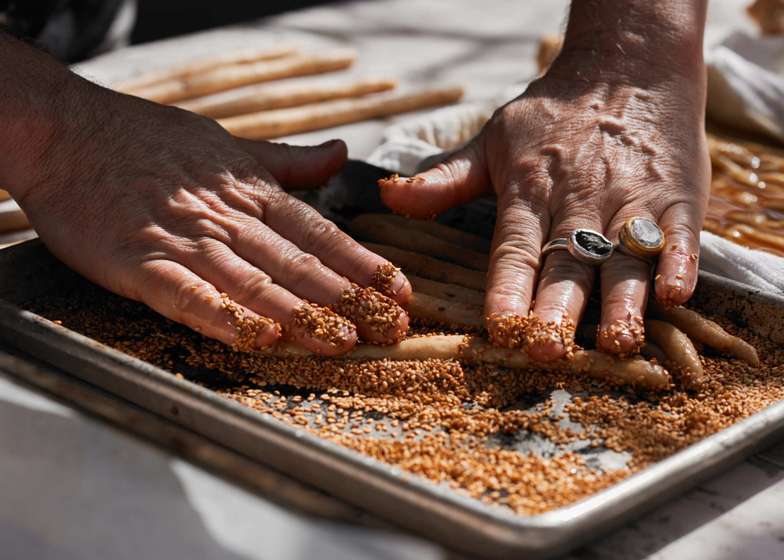 Hands with rinks atop baking tray with dough logs and sesame seeds