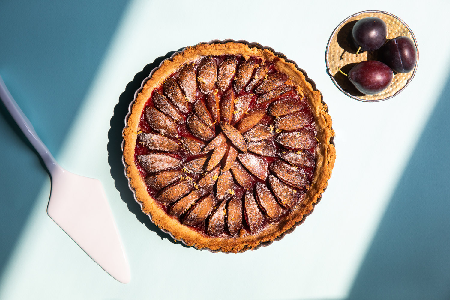 Plum tart on light blue background with small plate of plums next to it