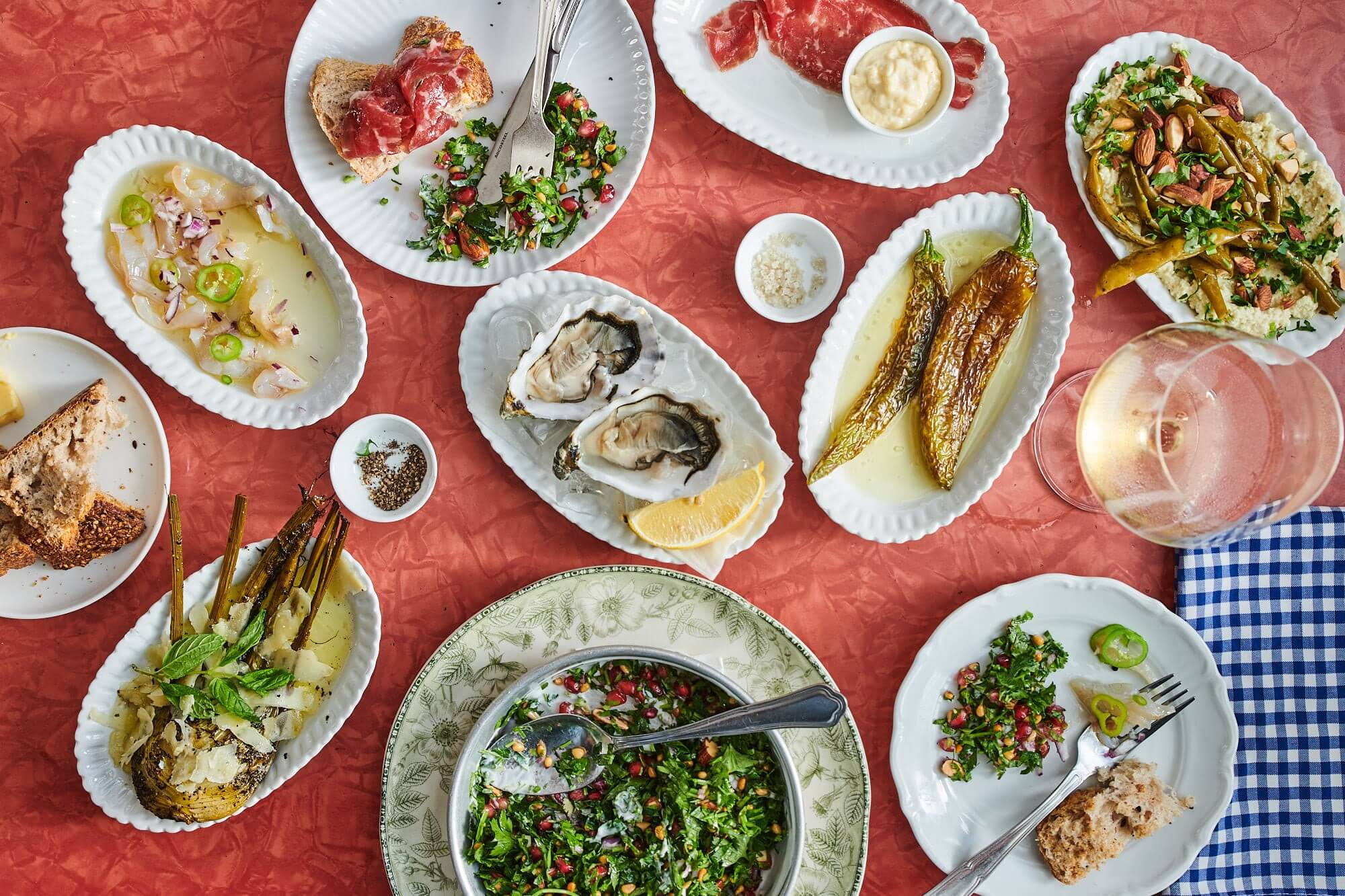 A red table is covered with lots of small white plates including oysters, cured meat, roasted peppers, and ceviche