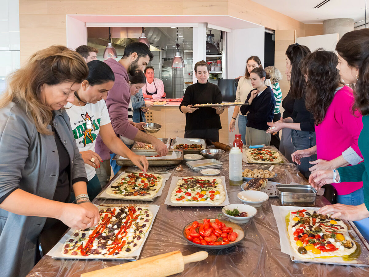A group of peope huddled around a long wooden table making focaccia topped with vibrant vegetables