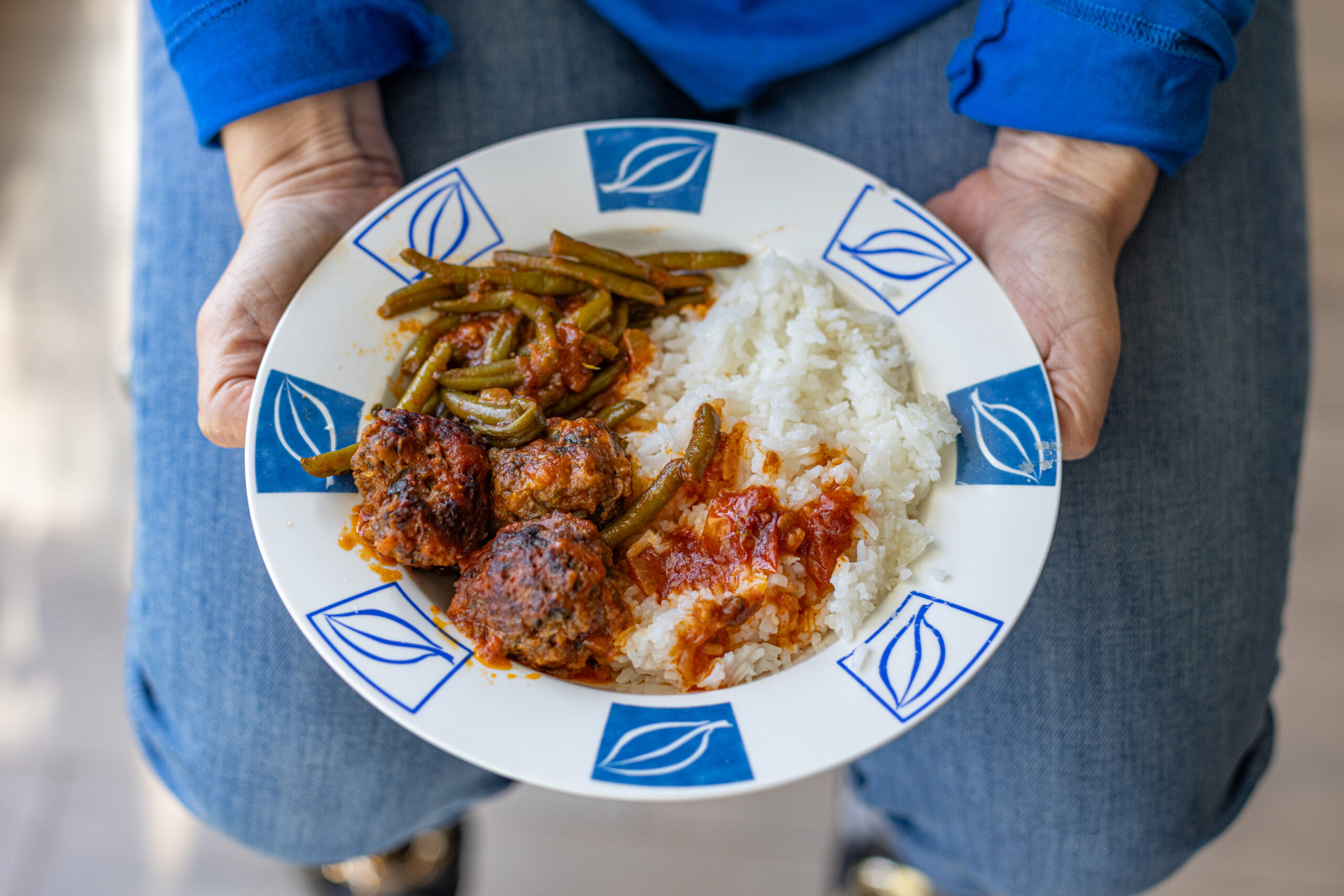 Person in jeans holding a blue and white plate of rice, green beans, and meatballs