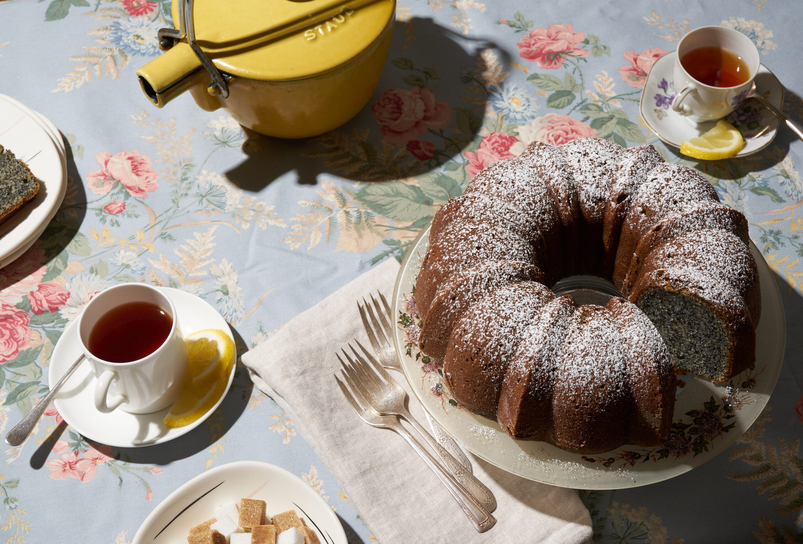Poppyseed bundt cake on a plate atop a blue tablecloth. Next to it are small glasses of tea with lemon slices.